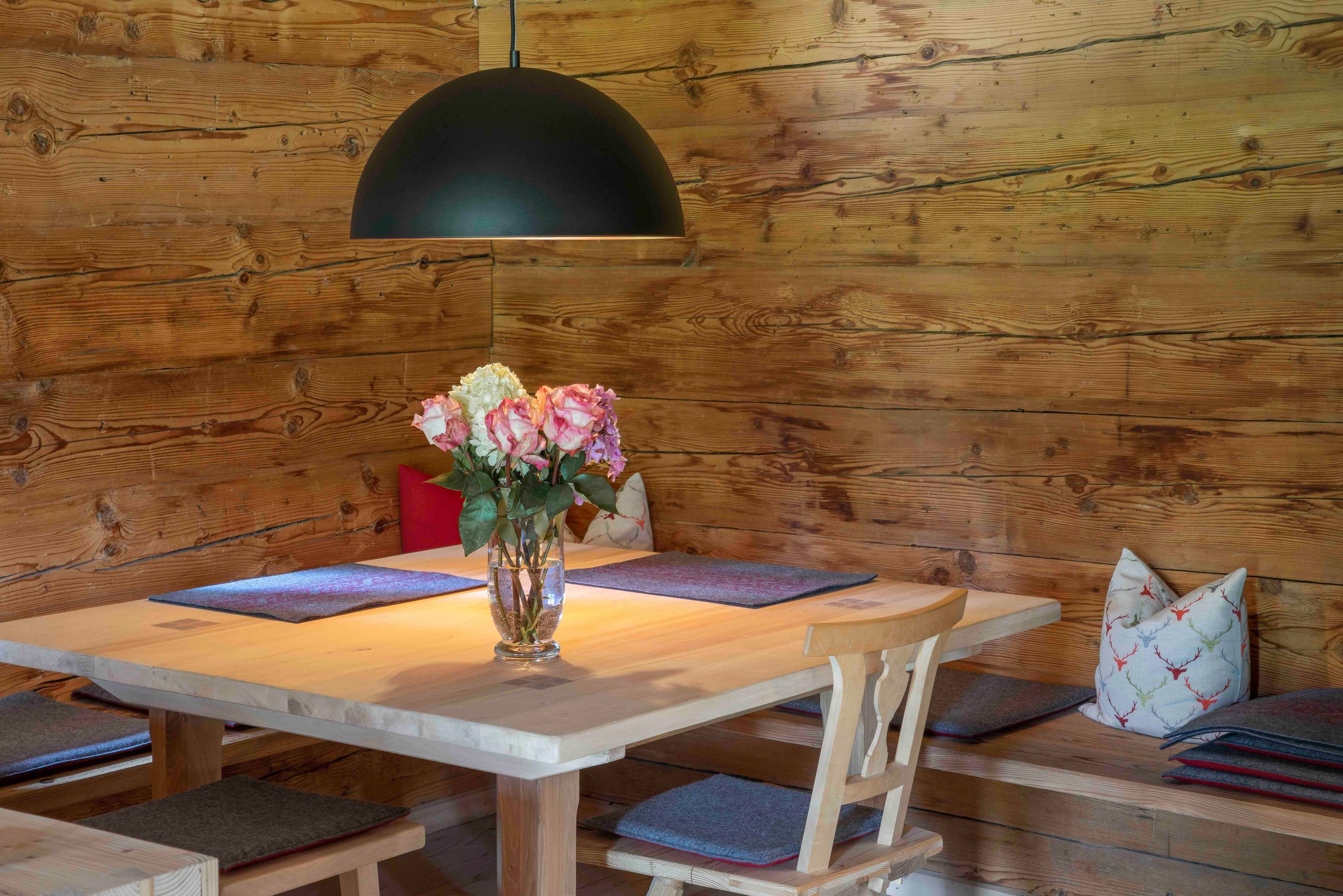 Holz-Essecke mit einem hellen Holztisch, Sitzbank, grauen Kissen, Blumen-Kissen, einer schwarzen Pendelleuchte und einer Vase mit rosa und weißen Rosen auf dem Tisch.