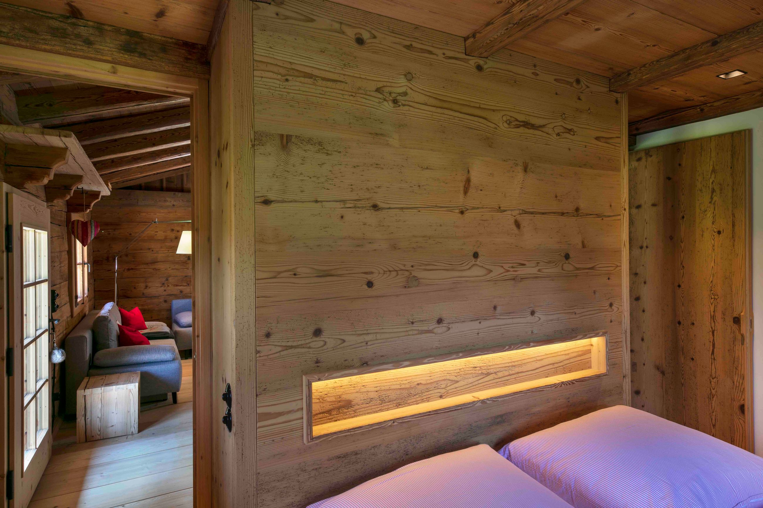 Gemütliches Schlafzimmer aus Holz mit zwei Betten, einem eingebauten, beleuchteten Regal in der holzgetäfelten Wand und einer Tür, die zu einer Sitzecke mit einem grauen Sofa und roten Kissen führt.