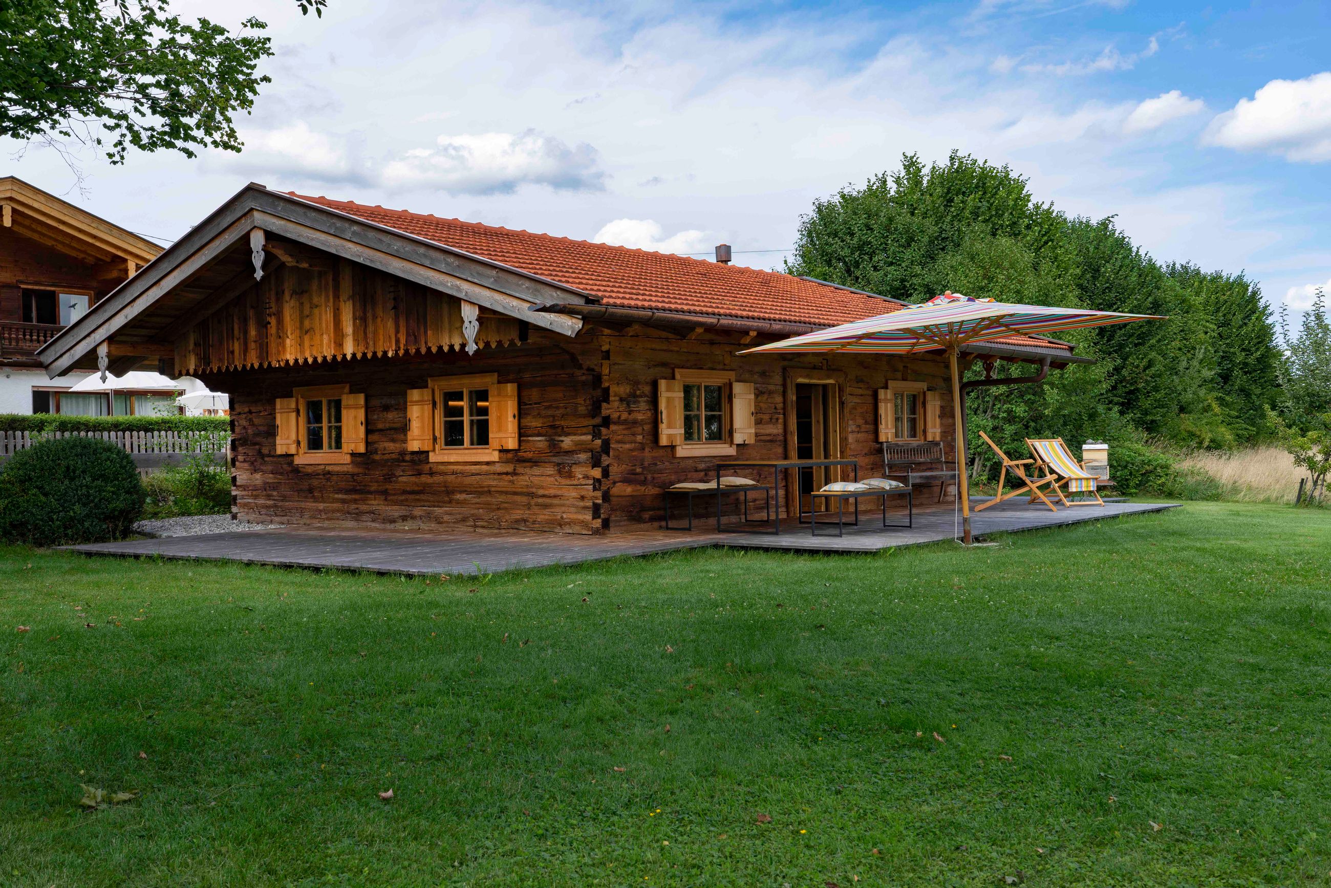 Rustikale Holzhütte mit rotem Schindeldach, kleinen Fenstern mit Fensterläden und einer Terrasse mit Sitzgelegenheiten im Freien unter einem großen gestreiften Sonnenschirm, umgeben von grünem Rasen und Bäumen.