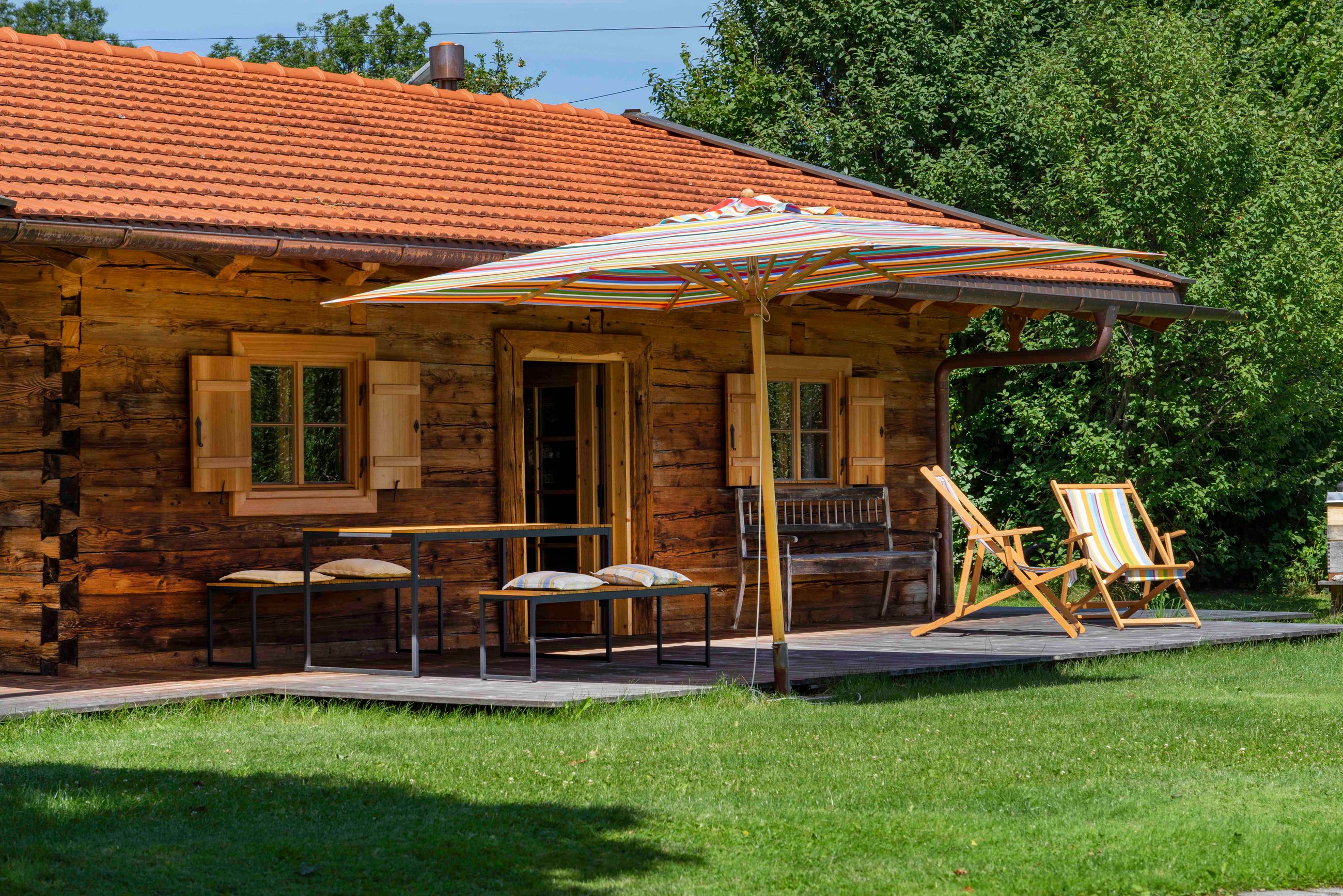 Holzhütte mit rotem Ziegeldach, zwei Fenstern und einer offenen Tür, einer Terrasse mit zwei gestreiften Strandstühlen, einer Bank, einem Tisch mit Kissen und einem großen bunten Sonnenschirm.